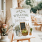 Homegrown baby shower welcome sign with a watering can, green apples, and baby overalls in sage green and neutral