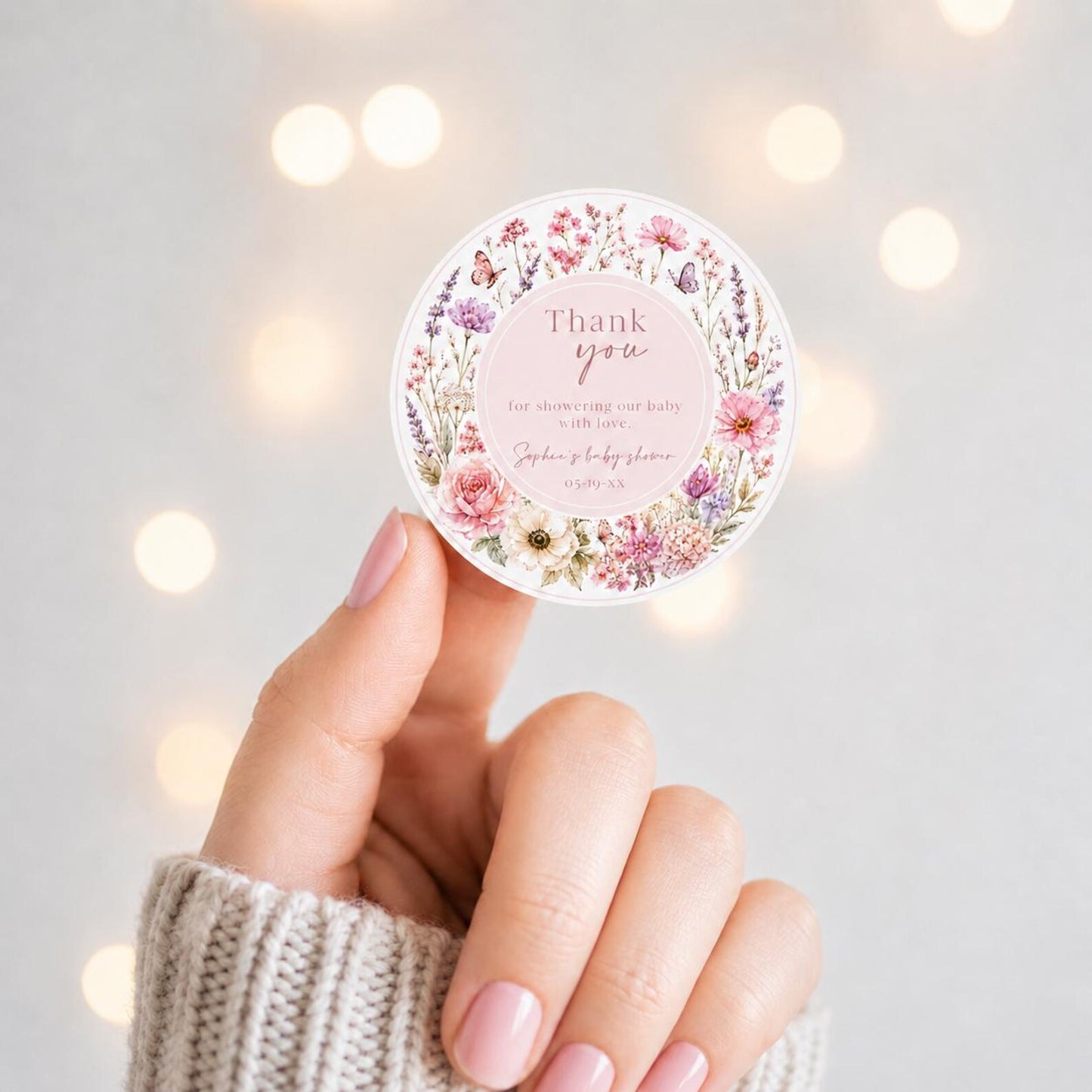 Editable wildflower baby shower round thank you sticker Canva template with watercolour roses, lavender, cosmos, butterfly details, and script typography on a soft blush background, shown tied to a kraft gift box