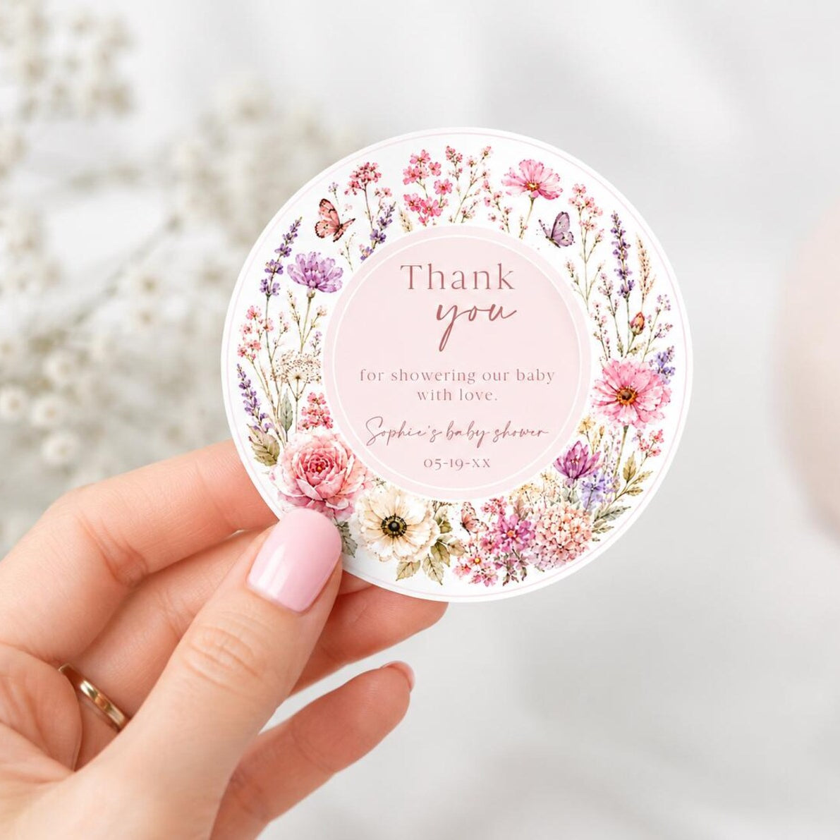 Editable wildflower baby shower round thank you sticker Canva template with watercolour roses, lavender, cosmos, butterfly details, and script typography on a soft blush background, shown tied to a kraft gift box